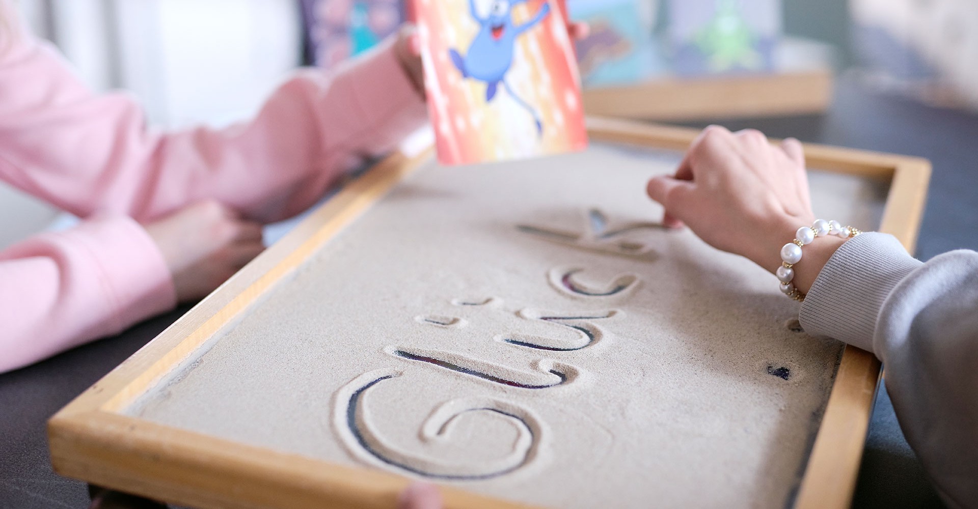 Zwei Kinder schreiben das Wort Glück in eine mit Sand gefüllte Kiste.