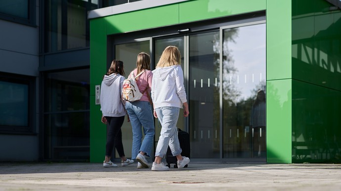 Das Bild zeigt drei junge Frauen, die nebeneinander herlaufen. Eine der Frauen schiebt einen Koffer.