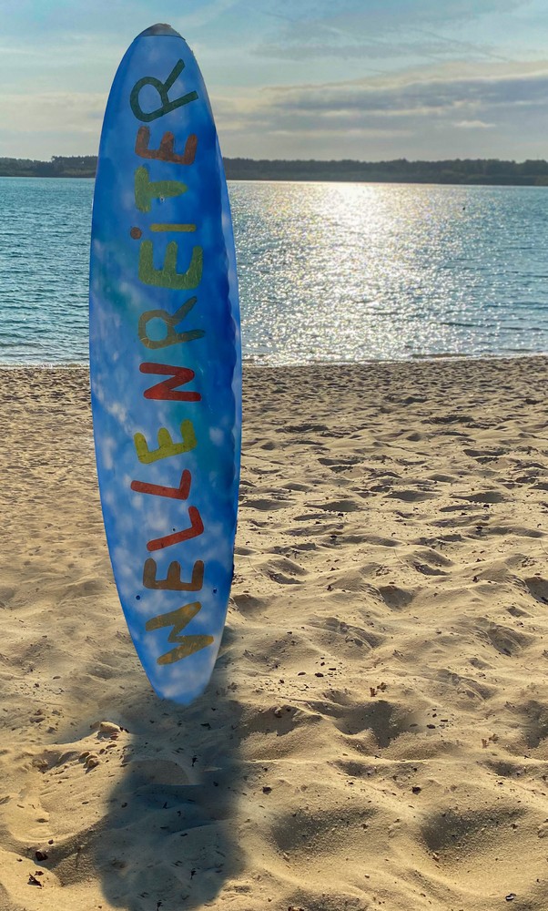 Das Bild zeigt ein Surfbrett an einem Strand, auf dem das Wort "Wellenreiter" steht.